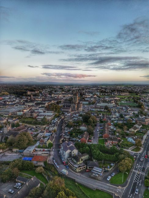 An expansive aerial view of Longford at dusk, showing a mix of residential streets, commercial buildings, and green spaces. In the foreground, a small park with neatly trimmed grass and a few trees is