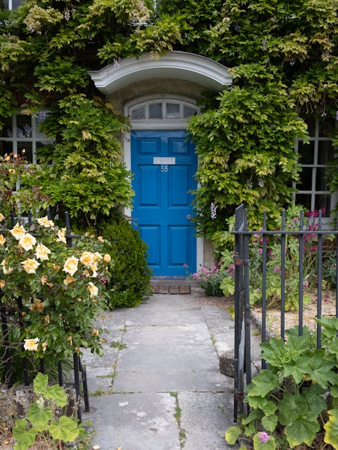 A charming garden pathway leads to a vibrant blue front door marked with the number 55, set within a white arched doorway surrounded by lush greenery. Overgrown ivy and leafy shrubs frame the entrance